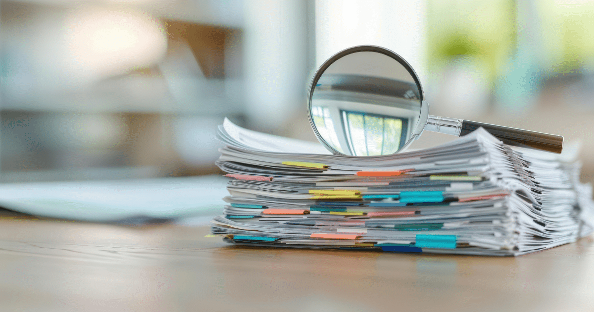 Stack of organized documents with colorful tabs and a magnifying glass on top, symbolizing a detailed review or audit.